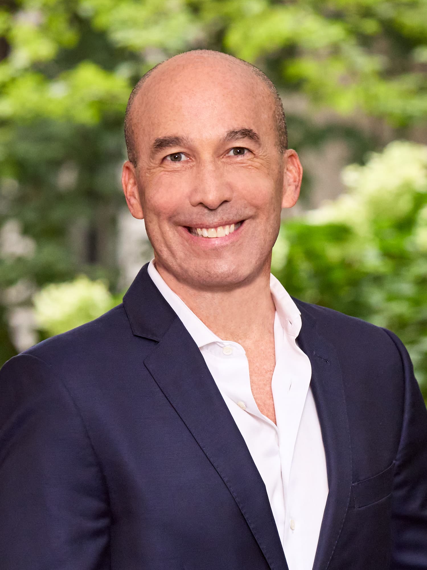 Smiling bald man in a navy blazer and white shirt with blurred green foliage.