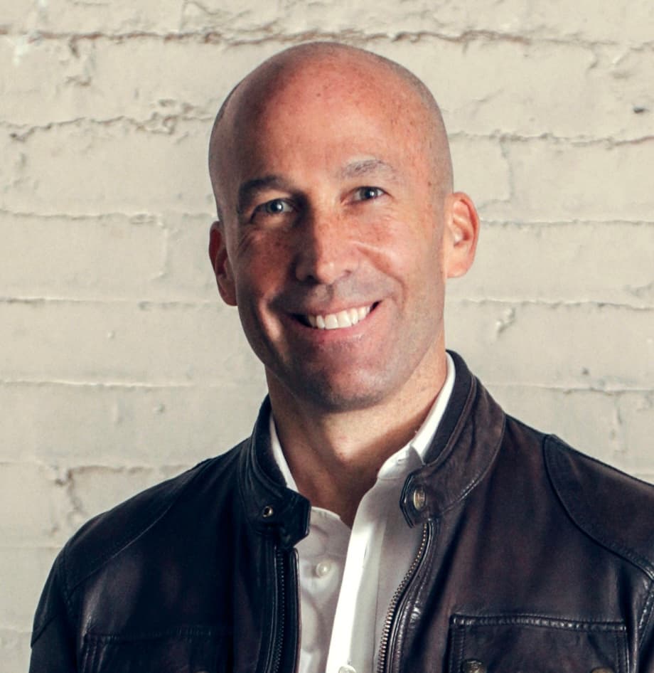 Smiling bald man in a brown leather jacket and white shirt against a brick wall.
